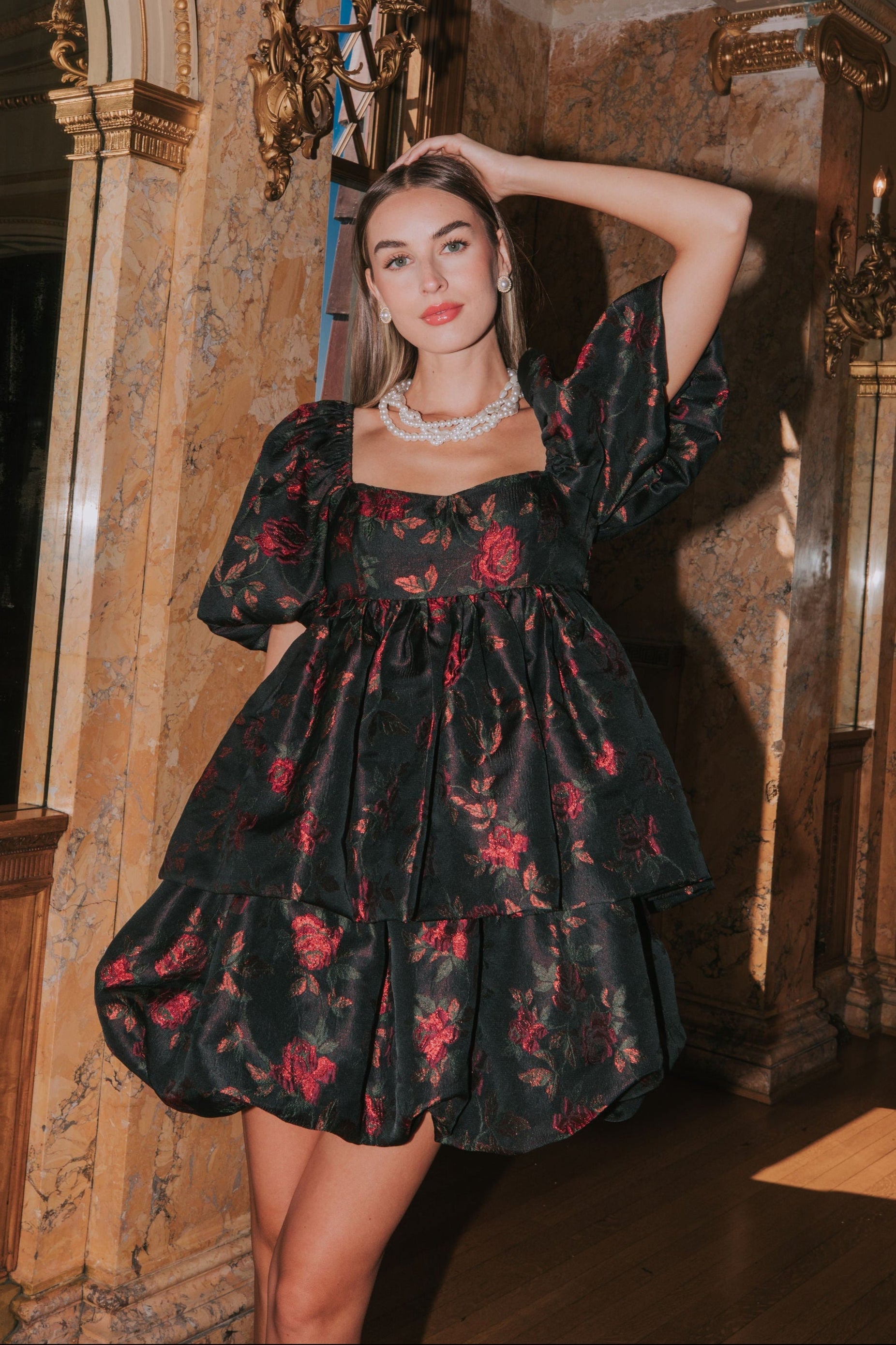 A woman wearing a black mini dress with red rose jacquard fabric, featuring a babydoll style with layered bubble skirt, sweetheart neckline, and bubble sleeves. She has a bow tie at the back and the dress appears to be fitted with elastic at the shoulders.