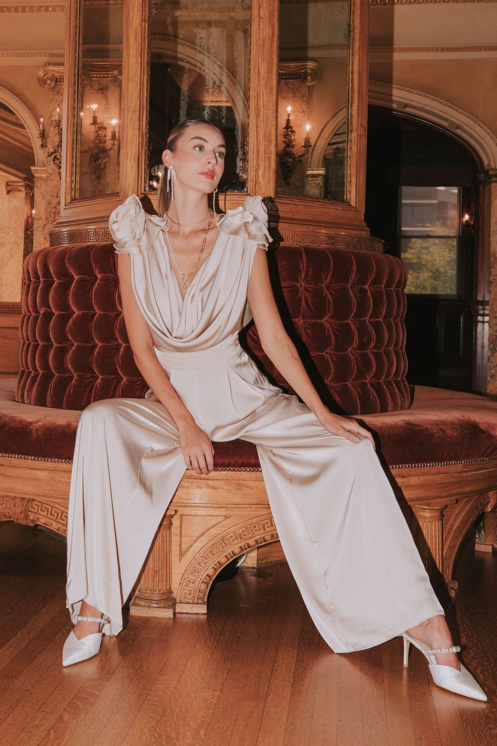 A woman sitting on a luxurious sofa wearing a champagne-colored sleeveless jumpsuit with a deep cowl neckline and 3D flowers on the shoulders. She has a tied bow at the back of her neck and is posing elegantly.