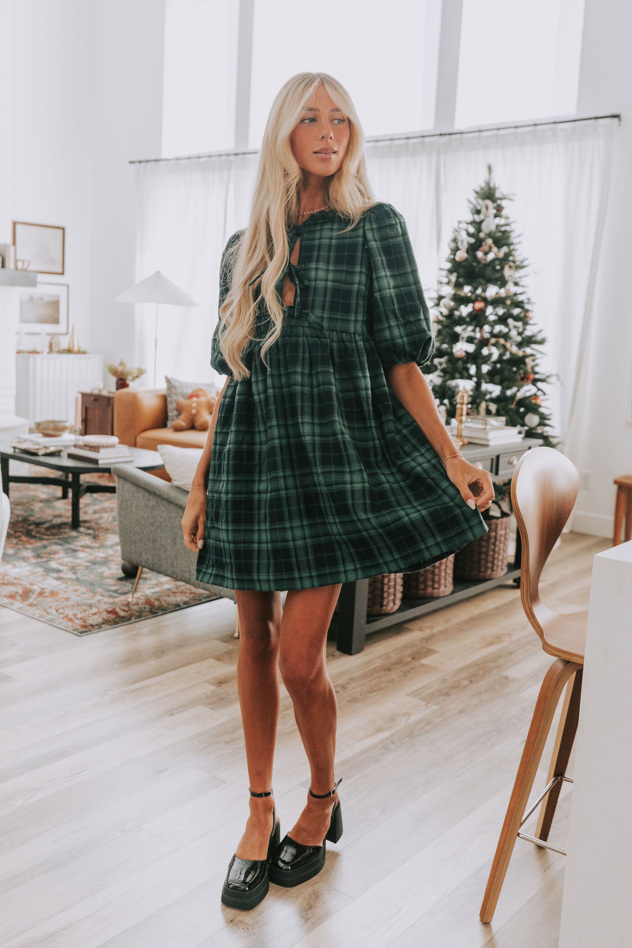 Woman in a green plaid dress standing in a modern living room with a Christmas tree.