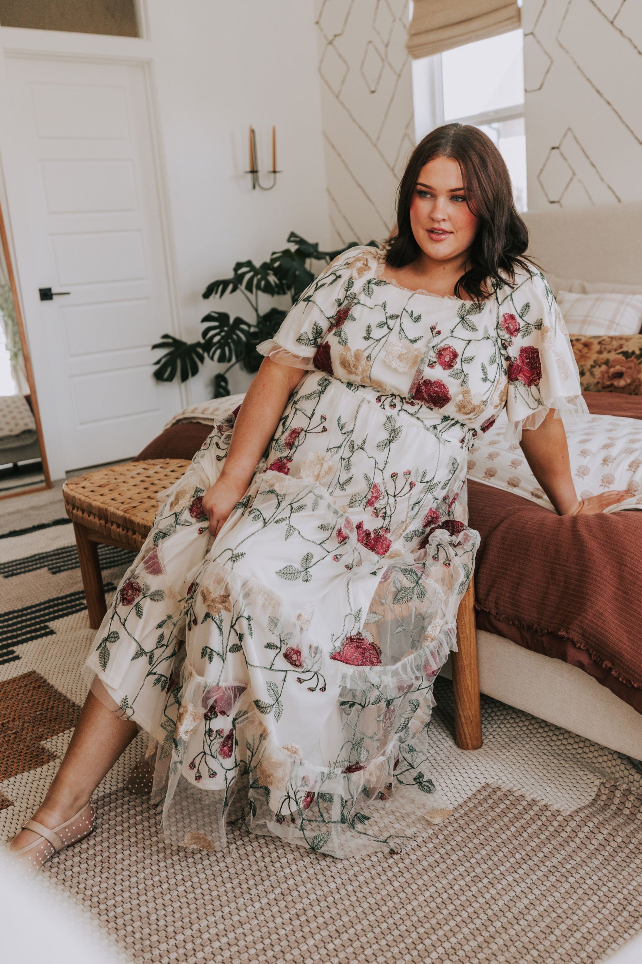 Woman in a floral dress sitting on a bed in a bedroom.