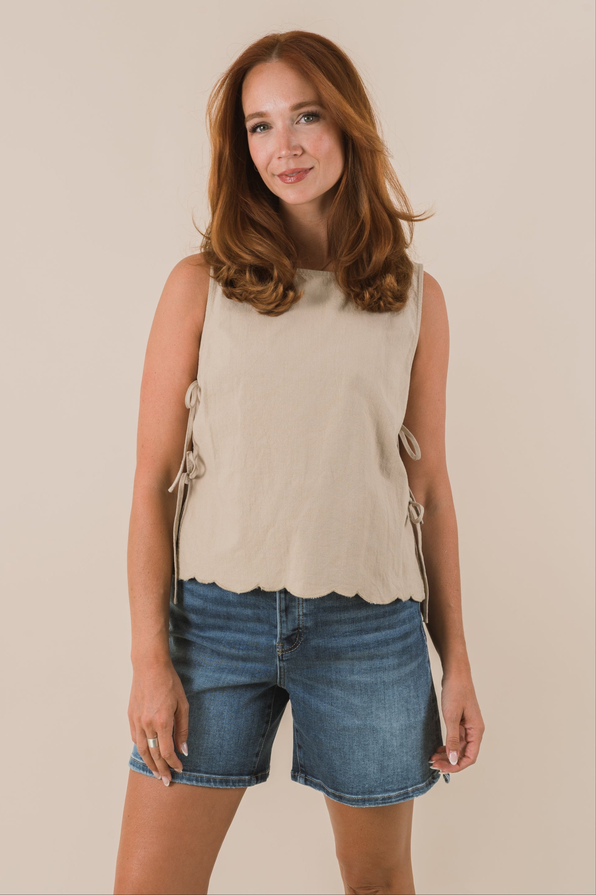 Woman wearing a beige sleeveless top with scallop details and blue shorts on a beige background