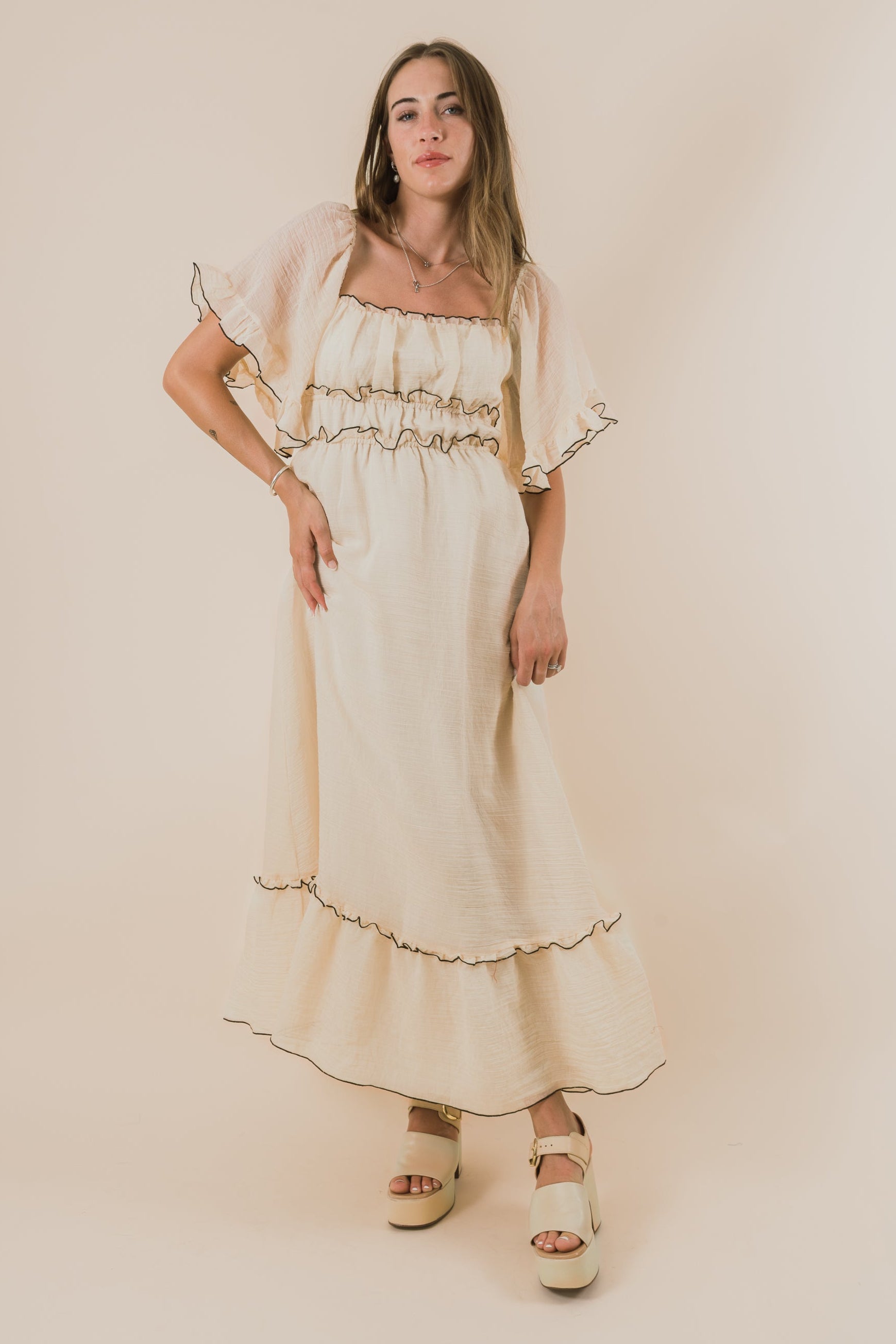 Woman wearing a long, light-colored dress with black ruffled accents against a plain background