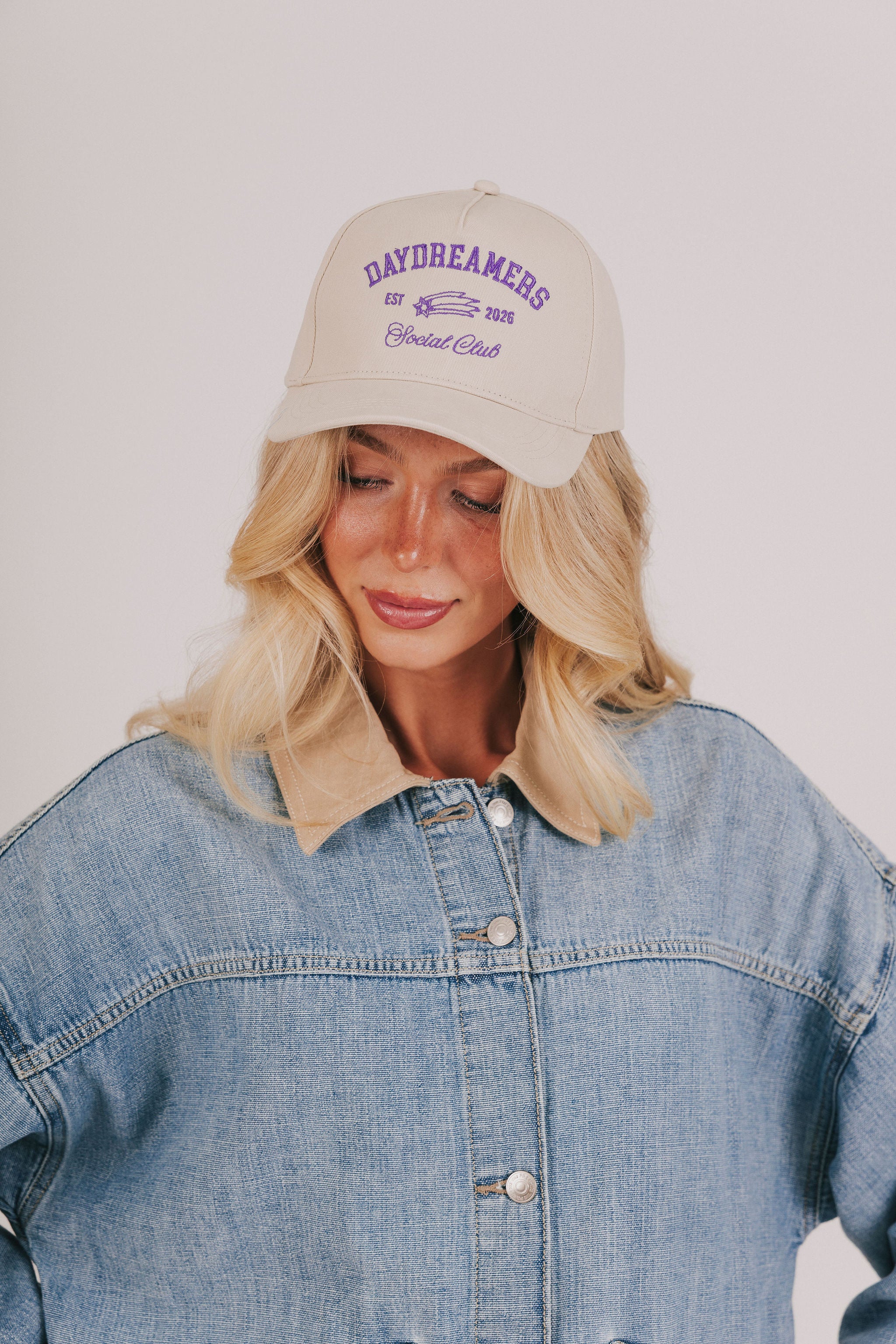 Woman wearing a beige cap with text and a denim jacket on a plain background