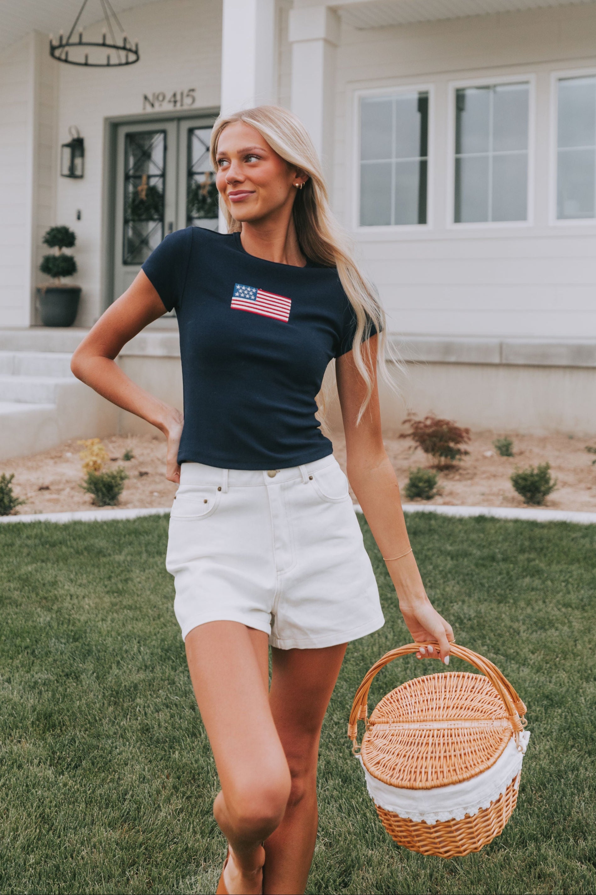 A woman wearing a dark navy T-shirt with an embroidered American flag, white denim shorts, and holding a wicker basket.