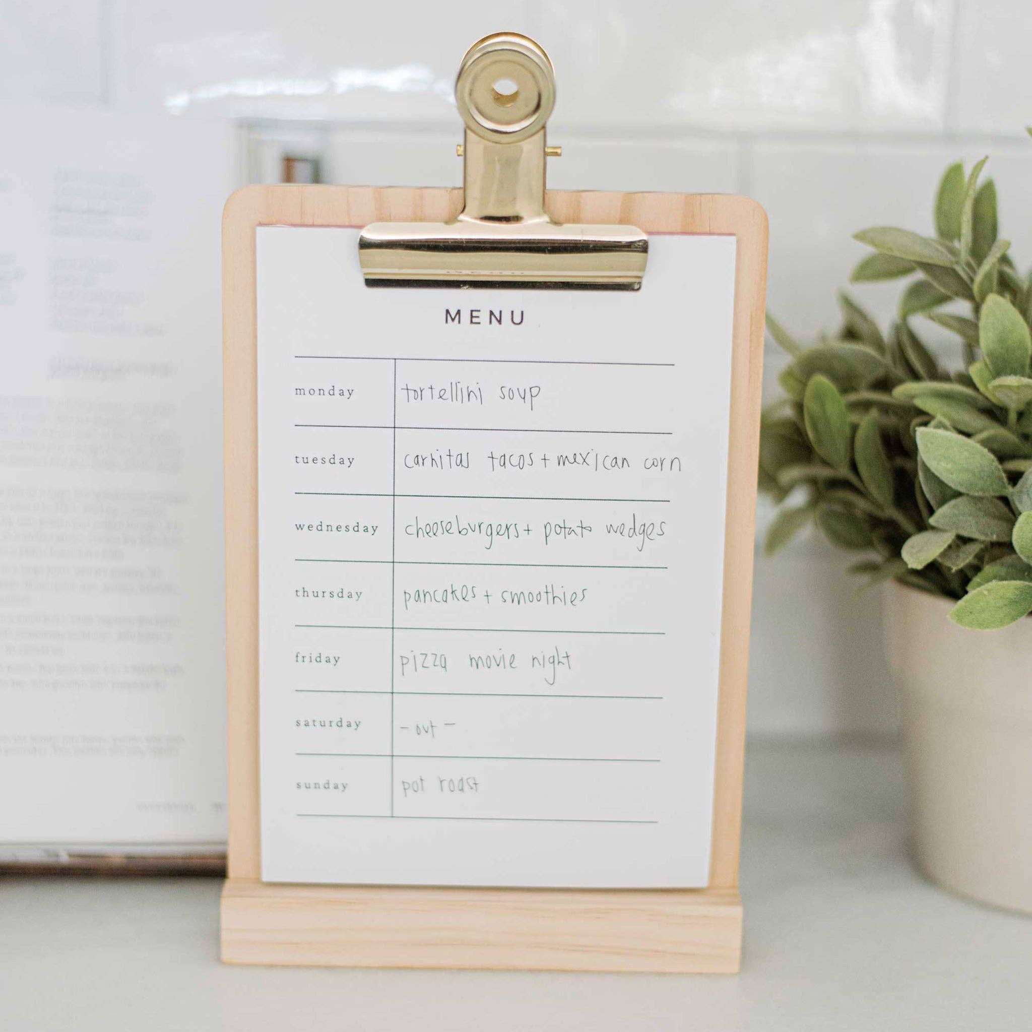 A wooden menu stand with a brass clip holding a paper notepad marked with each day of the week to plan a daily menu.