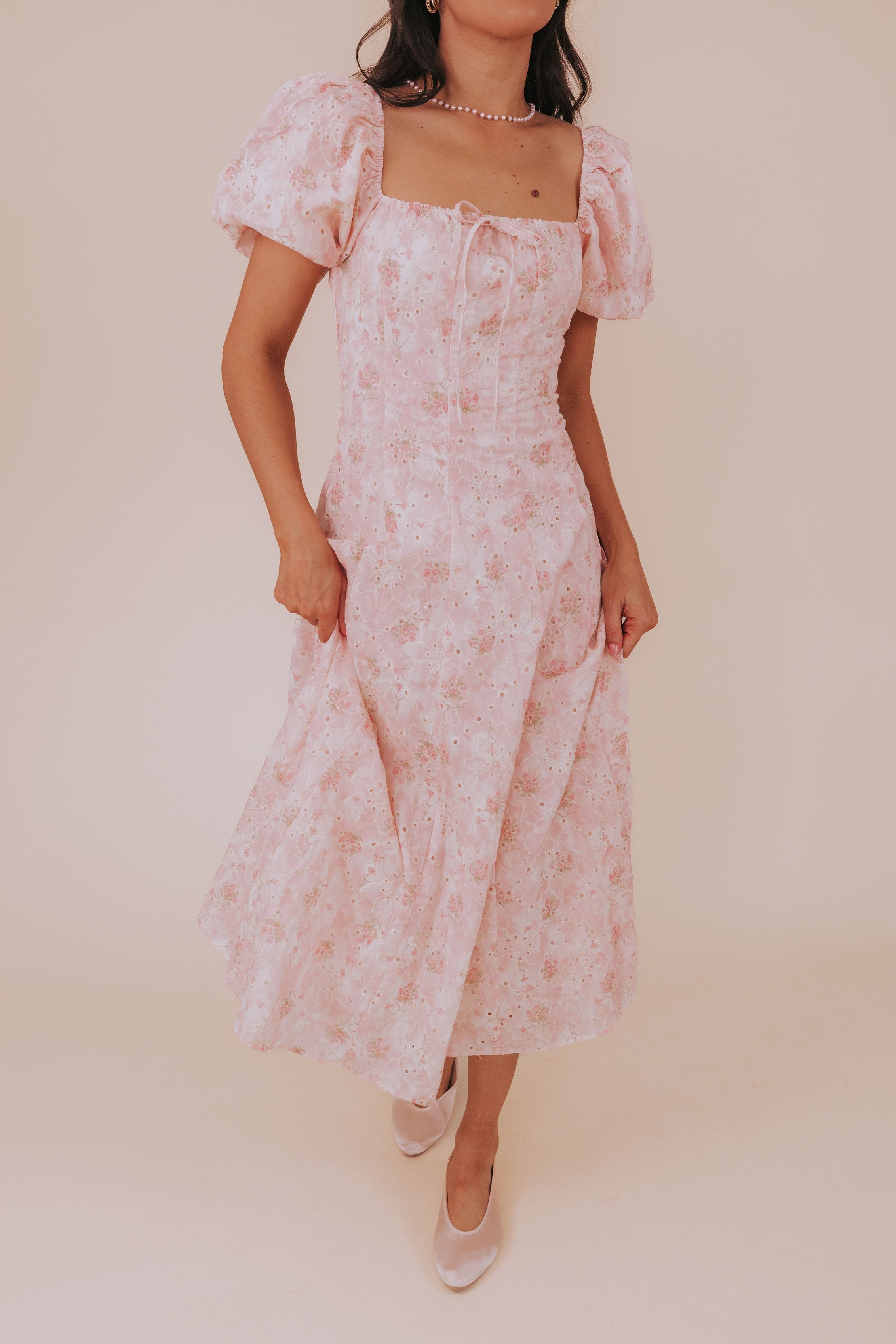 A woman wearing a pink floral dress with short bubble sleeves, square neckline with a decorative bow, and an invisible zipper in the back. The dress has cream eyelet embroidery and is midi length.