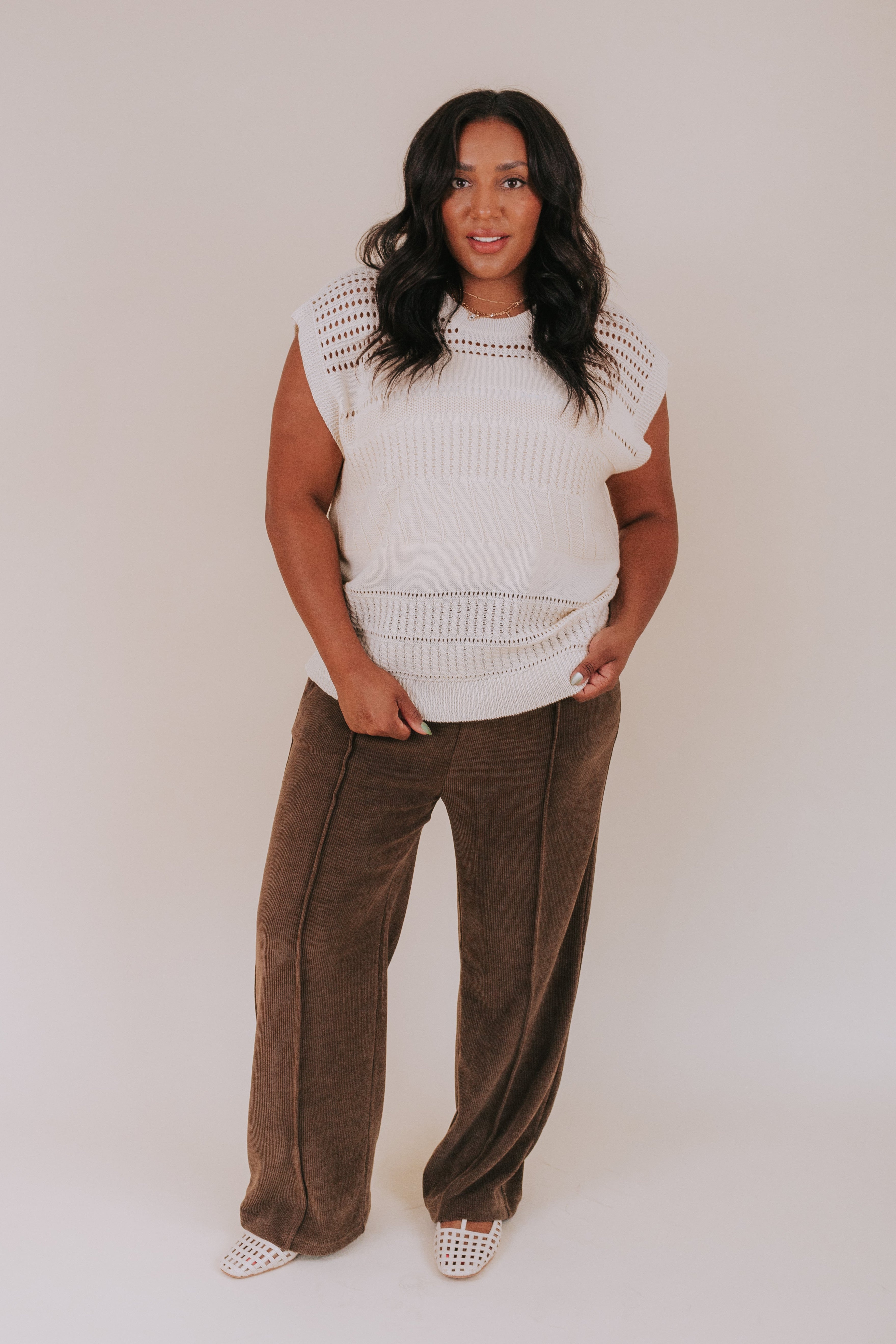 Woman wearing a white sleeveless sweater and brown pants against a plain background