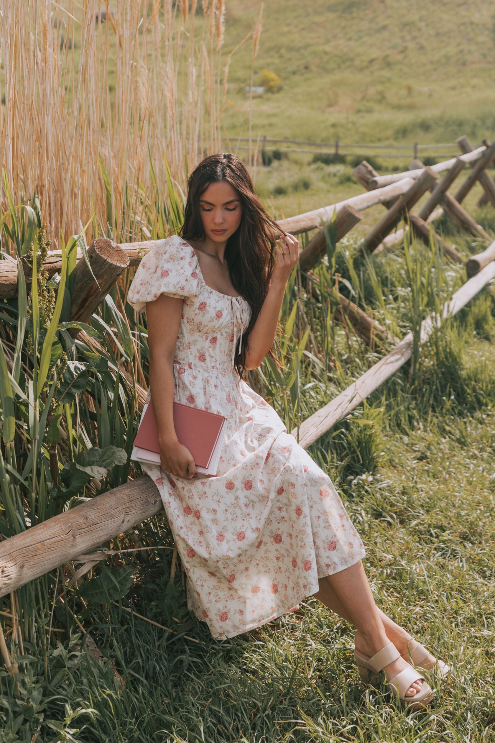 Woman in a floral dress sitting on a wooden fence in a rural setting with hills and corn stalks.