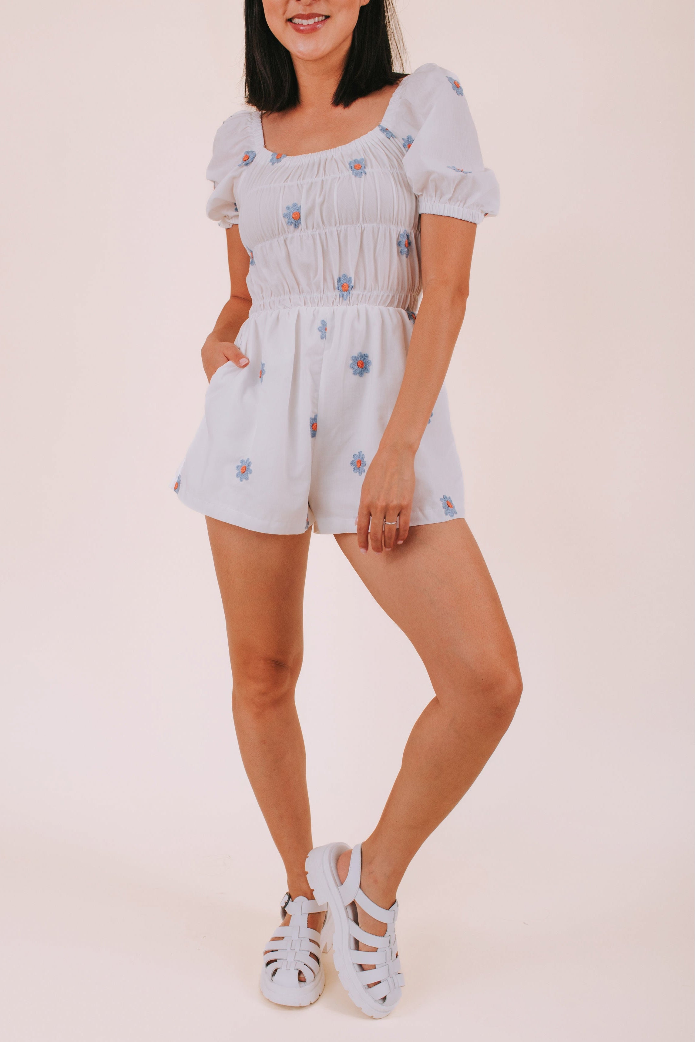 Woman wearing a white romper with blue daisies against a plain background