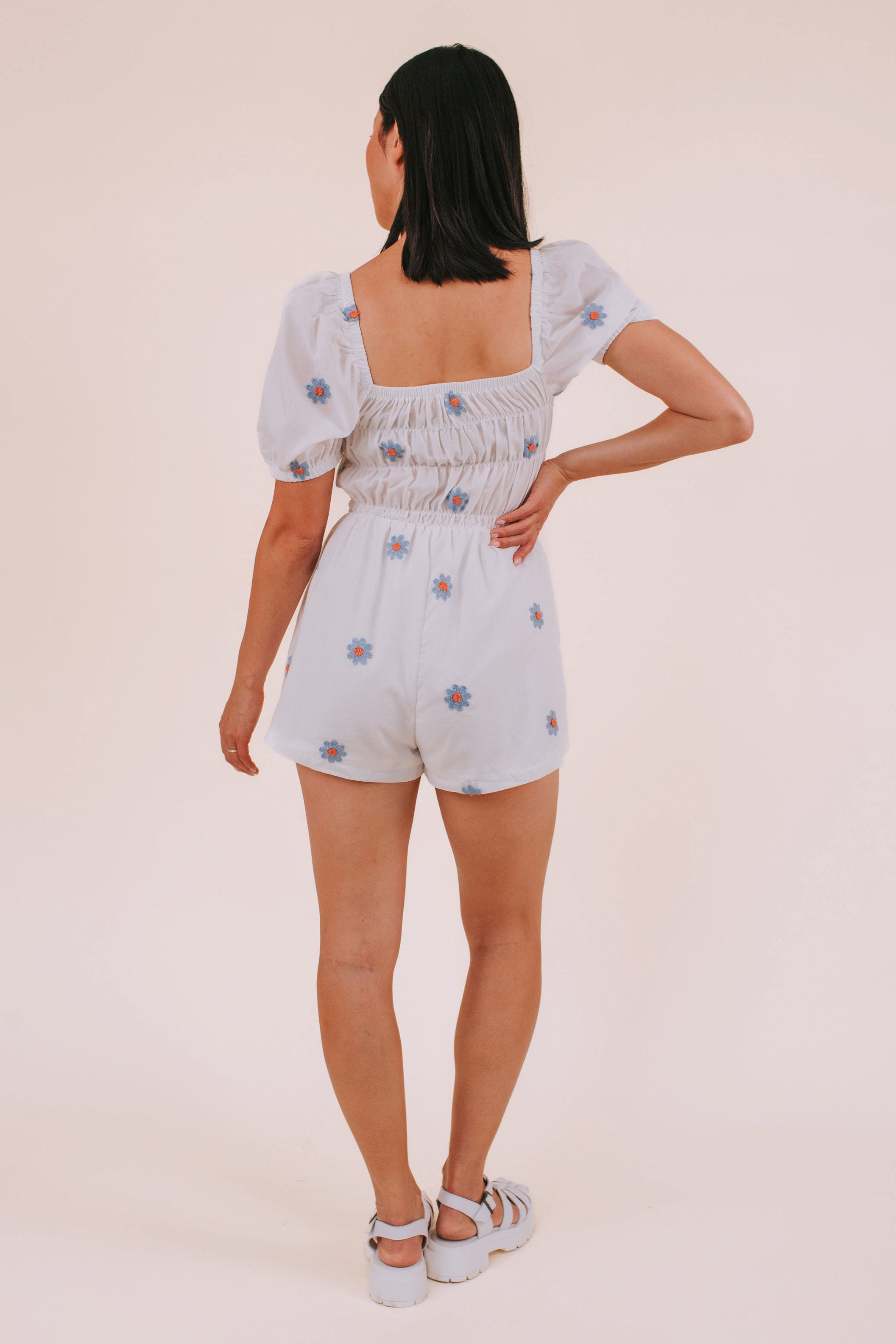 Woman wearing a white romper with blue daisies against a plain background