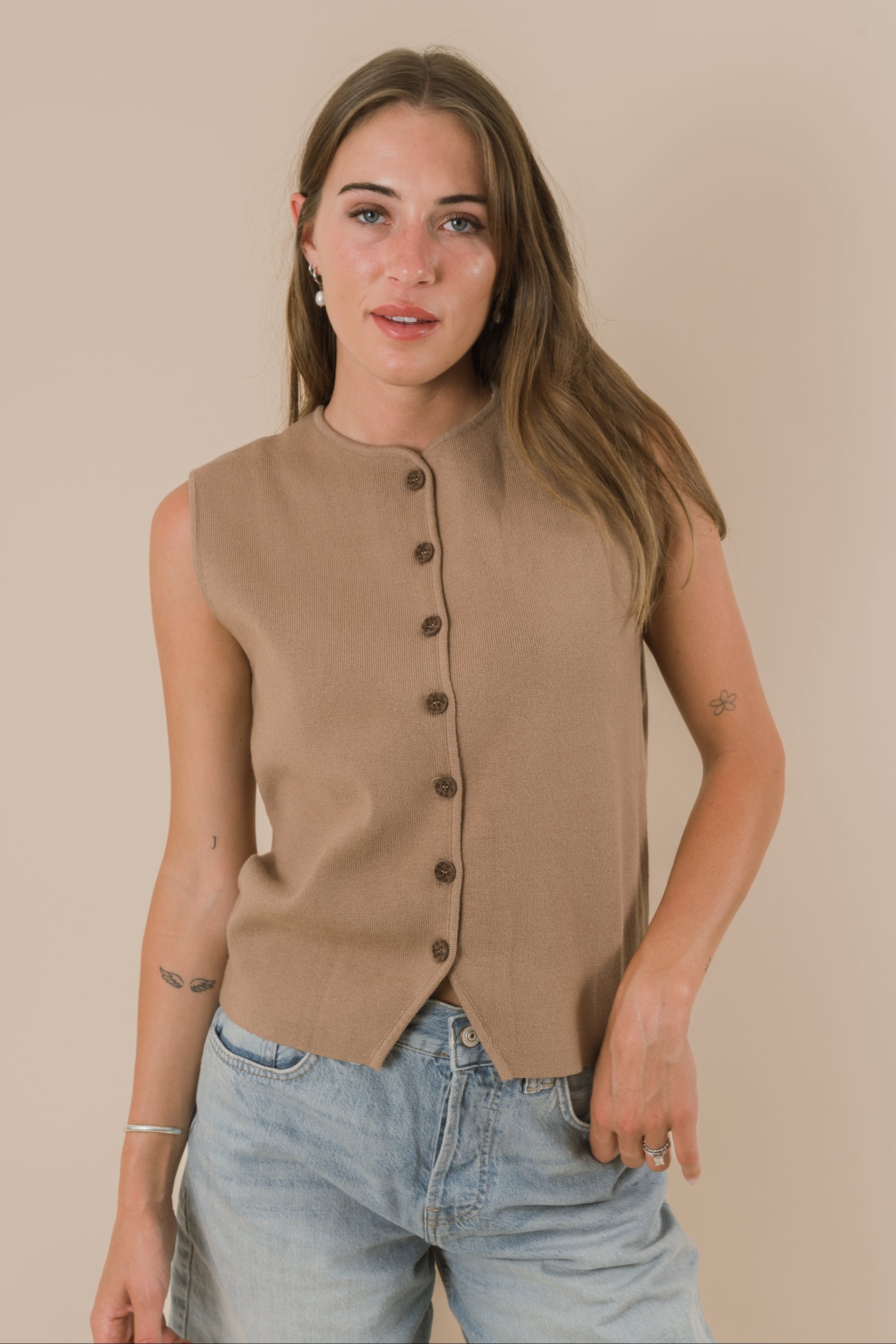 Woman wearing a beige sleeveless button-up top and light blue jeans against a beige background