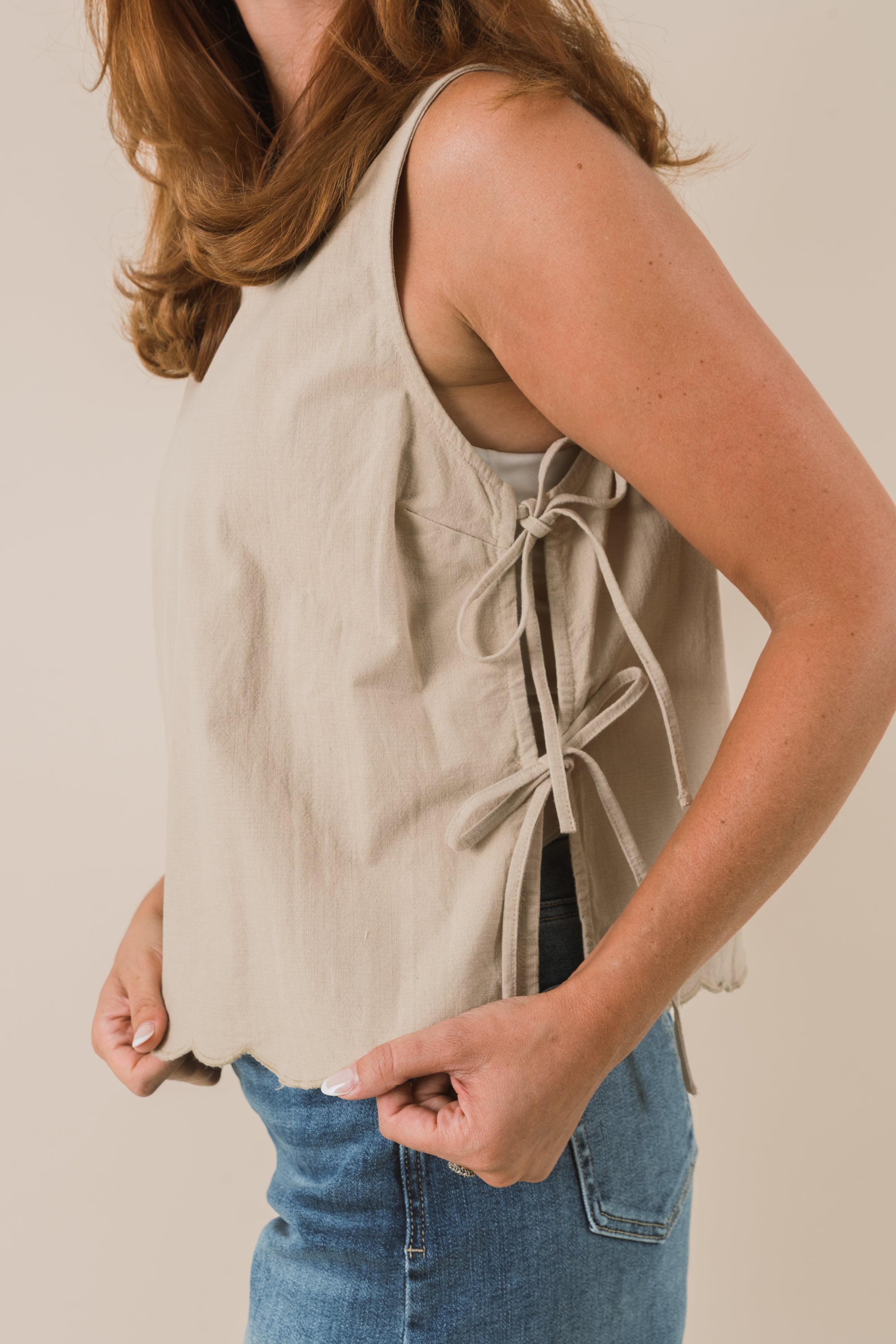 Person wearing a beige sleeveless top with tied bows on the side and blue jeans on a plain background
