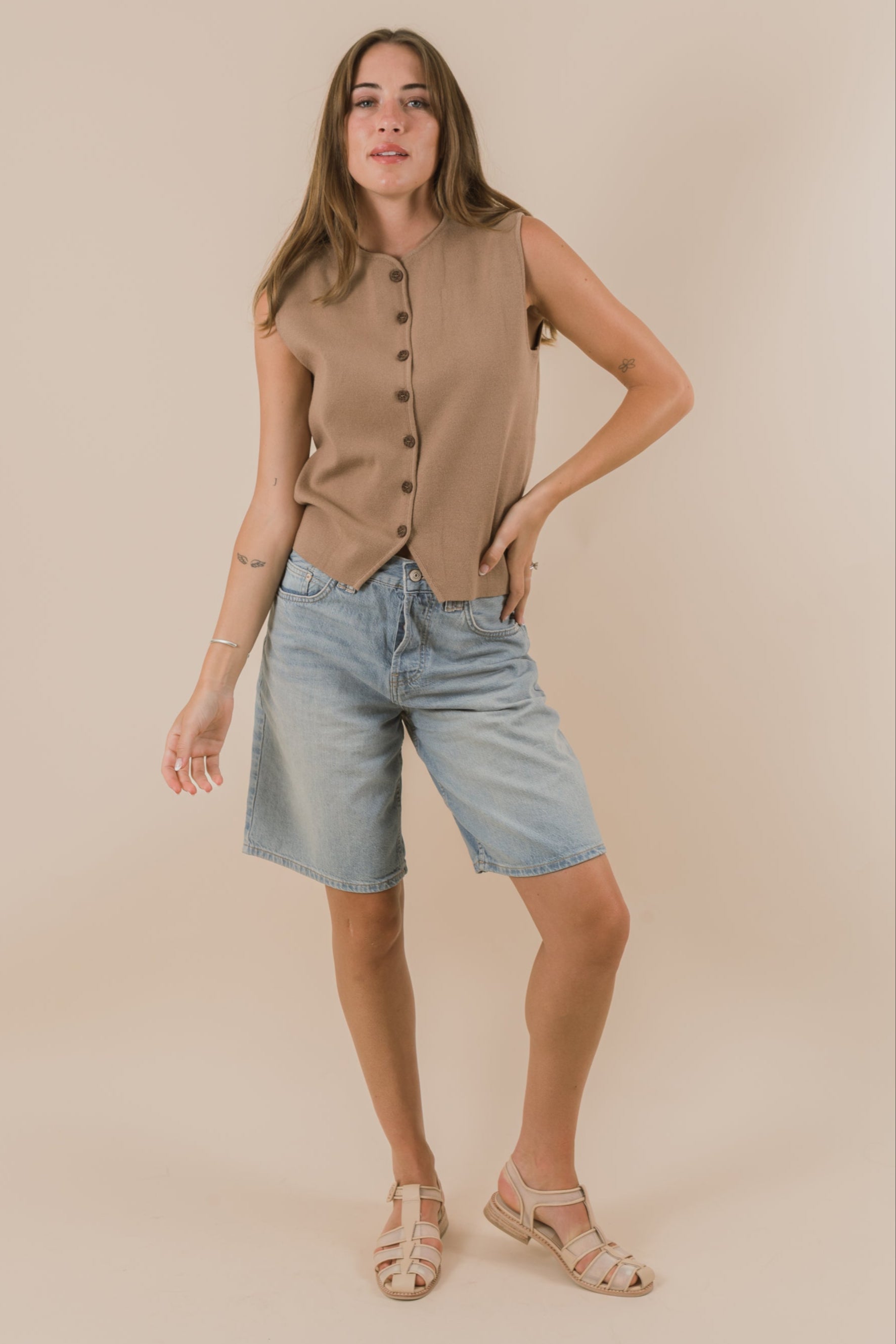 Woman wearing a beige sleeveless top and denim shorts on a beige background