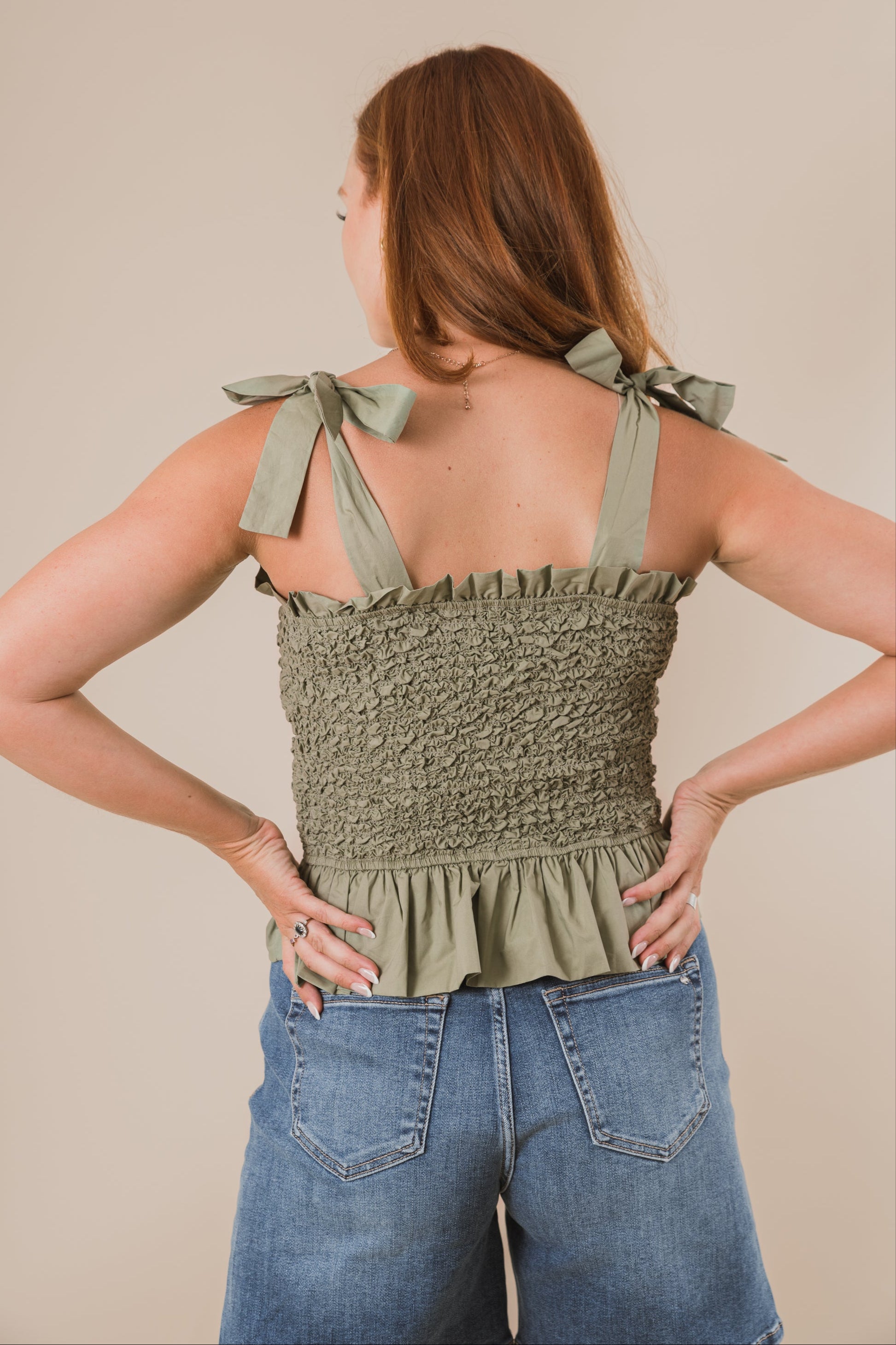 Woman wearing a green top with bow straps and blue jeans against a beige background