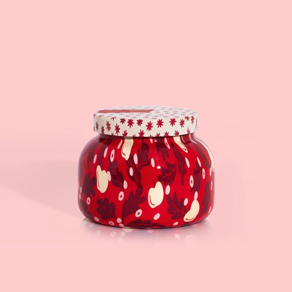 A red patterned jar with a white lid, adorned with images of apples and stars, containing a candle.