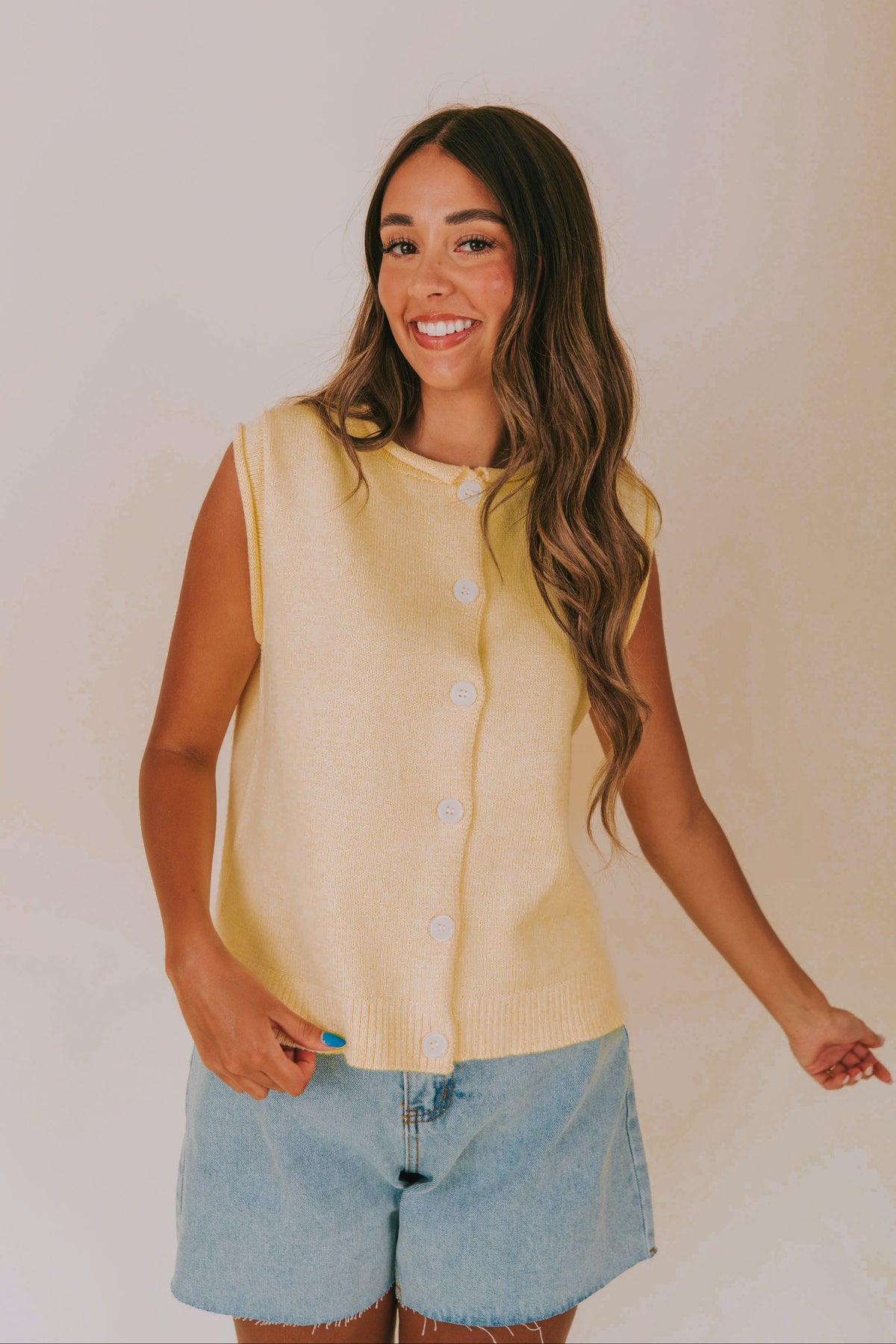 Woman wearing a yellow sleeveless cardigan and denim shorts against a plain background