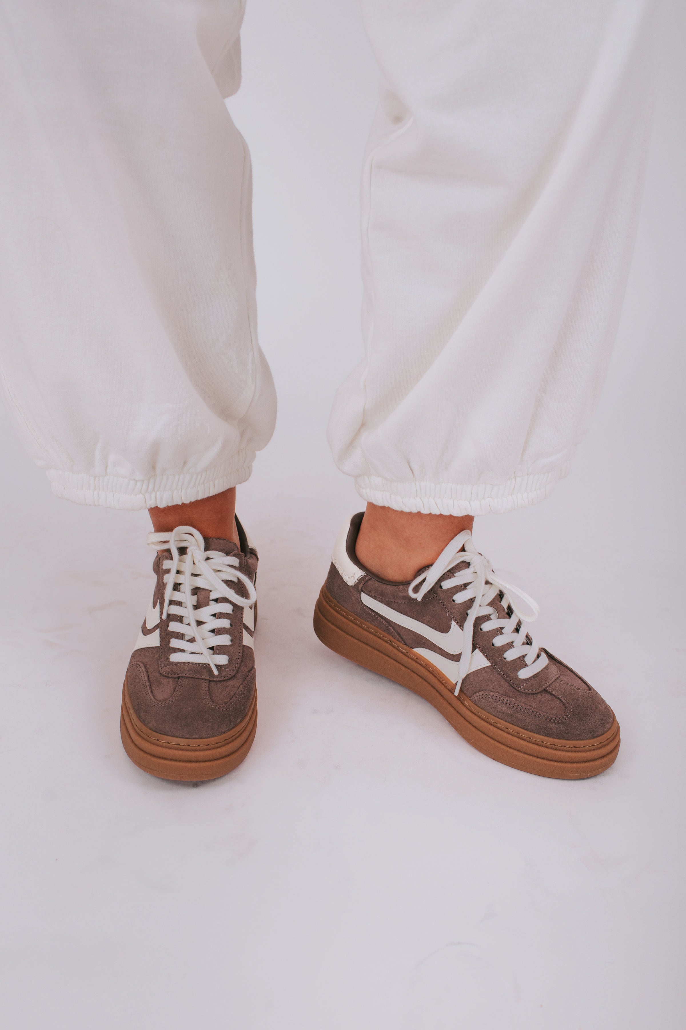 Brown sneakers with white laces worn with white pants on a light background