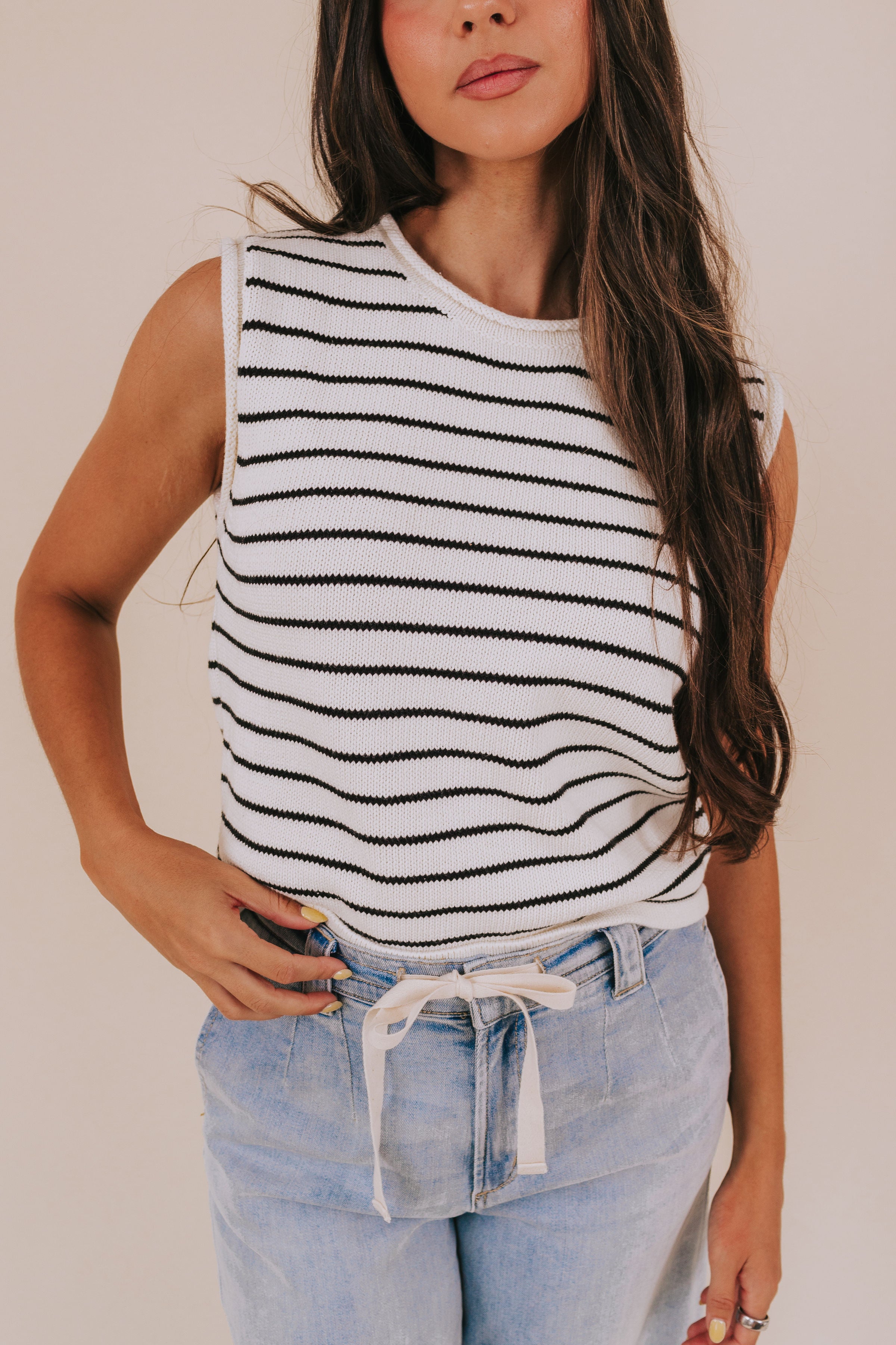 Woman wearing a black and white striped sleeveless top with light blue jeans against a plain background