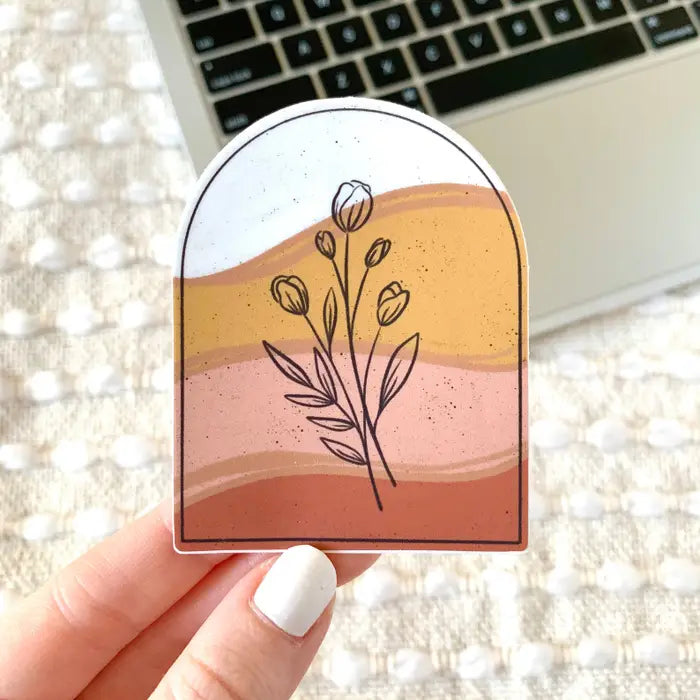 Hand holding a decorative sticker with floral design and color gradient in front of a laptop.