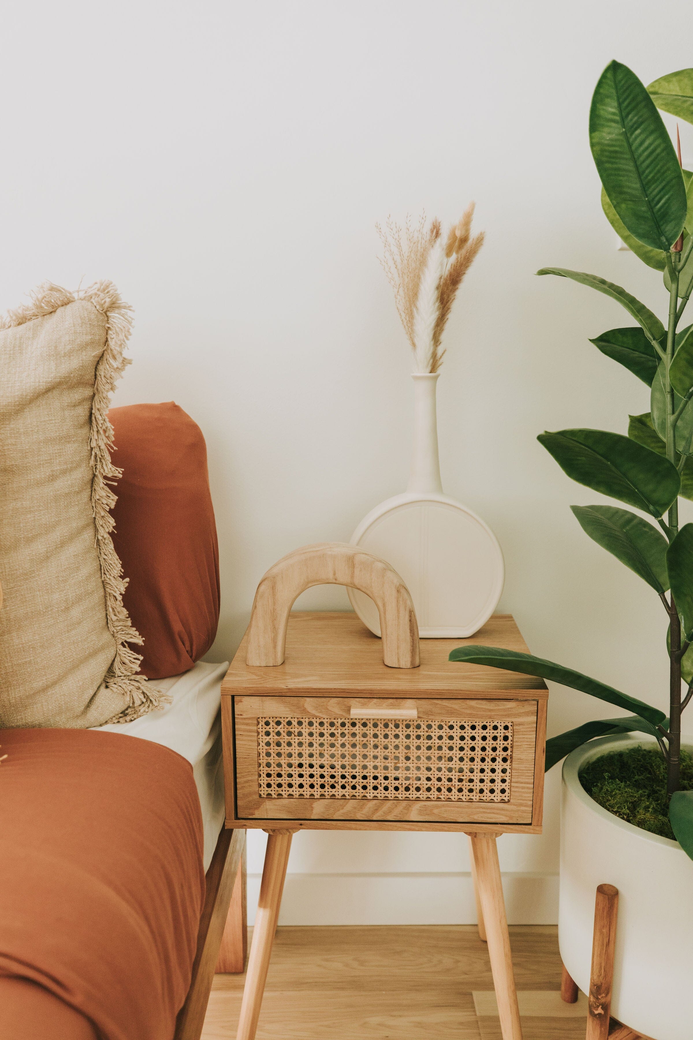 A white circular terracotta vase with a long neck placed on a wooden side table, surrounded by a cushion, a plant, and some decorative items.